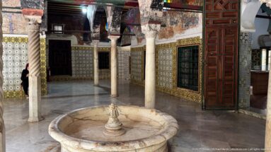Round stone fountain basin in a palace courtyard surrounded by slender columns, patterned tiles, windows, and a tall wooden door.
