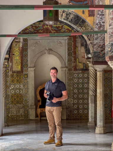 Man holding a camera beneath a palace arch, with spiral column, patterned tiles, painted ceiling, and weathered decoration.