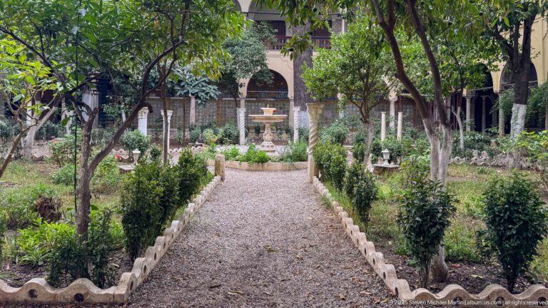 Gravel garden path bordered by clipped shrubs and mature trees leading toward a small fountain and palace arcades.