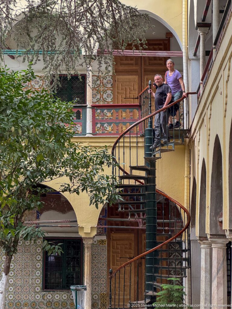 Two men standing at the upper landing of a dark metal spiral staircase amid tiled walls, painted doors, arcades, and foliage.