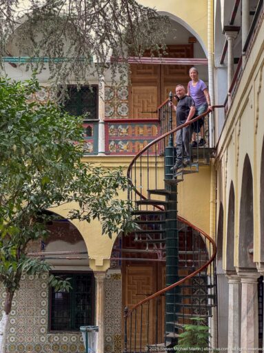 Two men standing at the upper landing of a dark metal spiral staircase amid tiled walls, painted doors, arcades, and foliage.