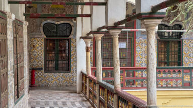 Upper palace gallery with patterned tile walls, slender stone columns, dark windows, painted railing, and black-and-white floor.