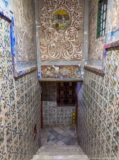 Descending palace passage lined with floral tiles beneath an ornate painted ceiling with central medallion and scrolling decoration.