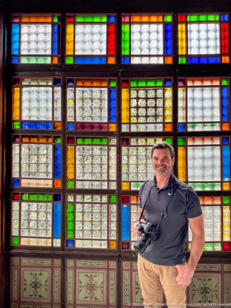 Man holding a camera standing before a grid of red, blue, green, amber, and patterned glass windows.