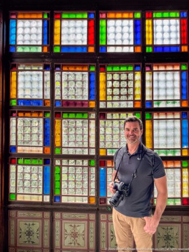 Man holding a camera standing before a grid of red, blue, green, amber, and patterned glass windows.