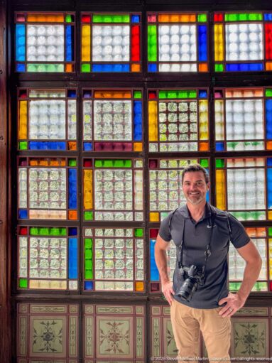 Man with camera around his neck posing hands-on-hips before multicolored geometric glass windows.