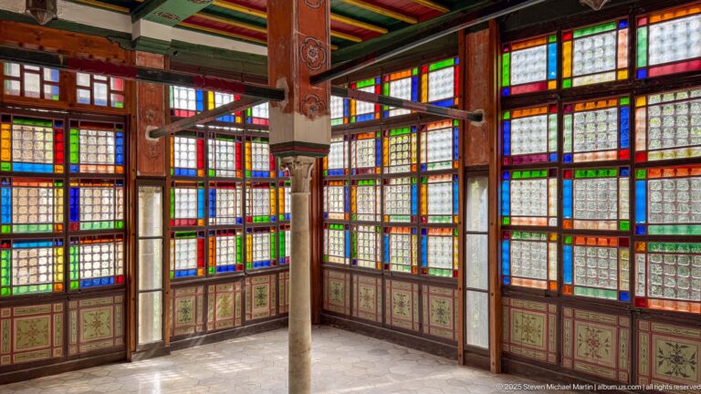 Interior corner with tall multicolored glass windows, painted lower panels, slender columns, and brightly painted ceiling beams.