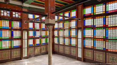 Interior corner with tall multicolored glass windows, painted lower panels, slender columns, and brightly painted ceiling beams.