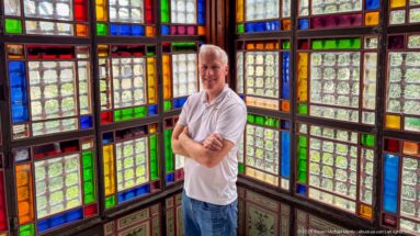 Man in a white shirt with folded arms standing in a corner surrounded by multicolored patterned glass windows.