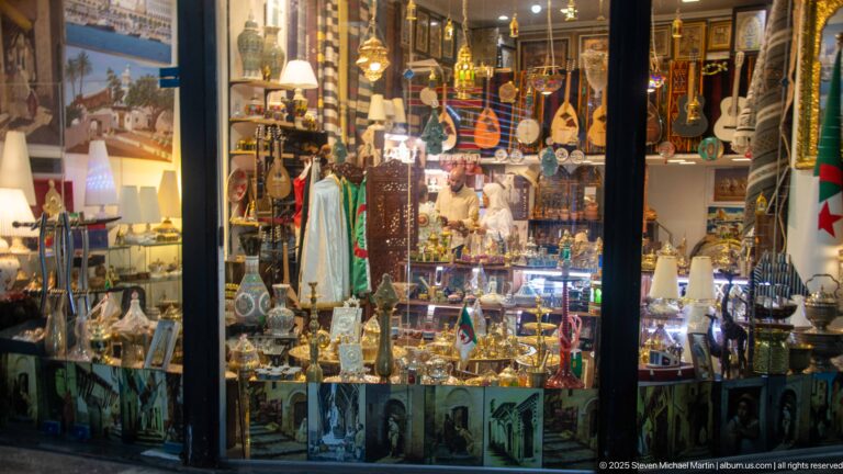 Souvenir shop window filled with stringed instruments, metalware, textiles, lamps, and small Algerian flags.