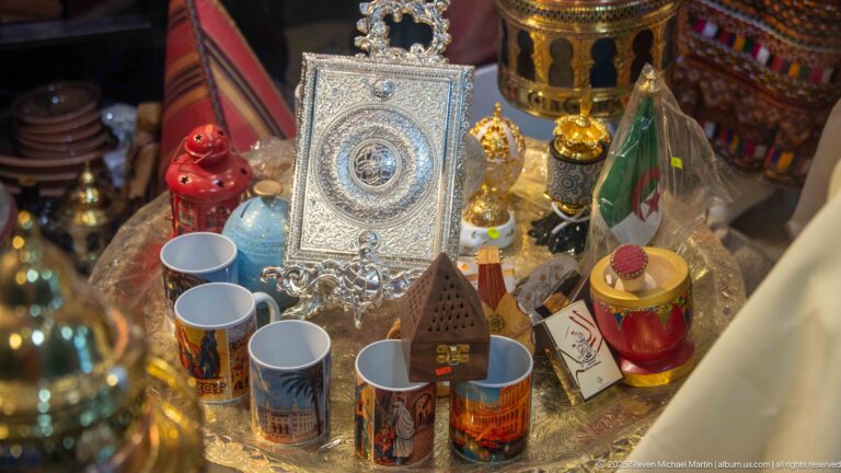 Souvenir display with an Algerian flag, mugs, ceramics, incense burners, and ornate silver-colored metalwork.