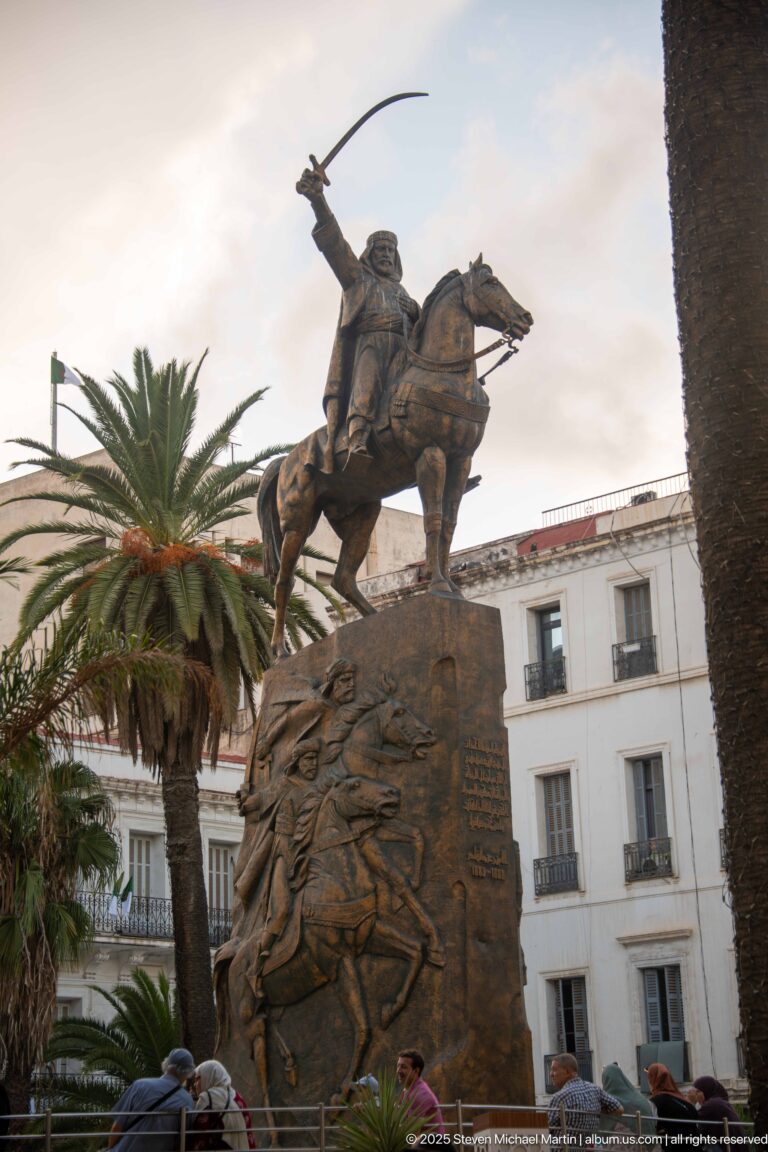 Side view of a bronze-colored rider raising a sword on horseback above a relief-covered pedestal.