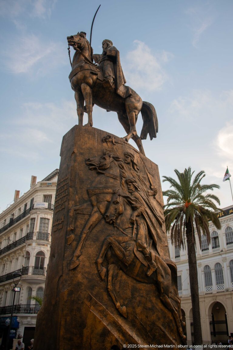Bronze-colored equestrian statue of a sword-wielding rider on a tall relief-covered pedestal with palms and buildings behind.