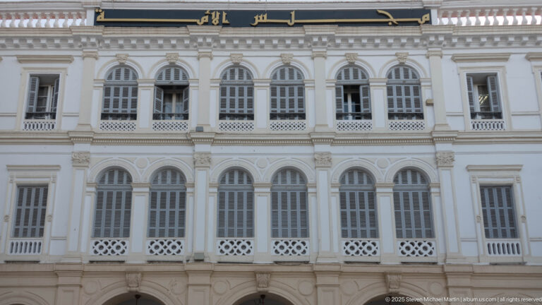 White symmetrical façade with repeated arched windows, decorative pilasters, lattice panels, and Arabic lettering near the roofline.