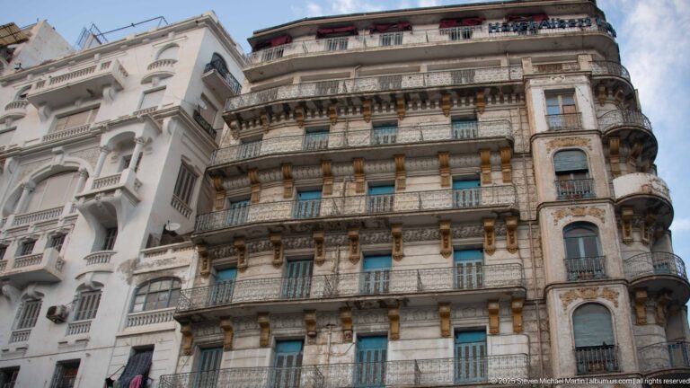 Adjacent multi-story buildings with white and stone façades, projecting balconies, green shutters, and ornate detailing.