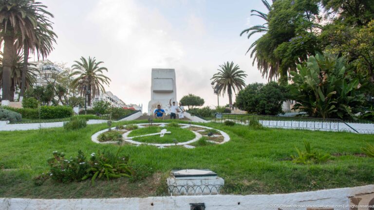 Formal green lawn with circular flowerbed, palms, clipped trees, and a tall pale monument on the central axis.