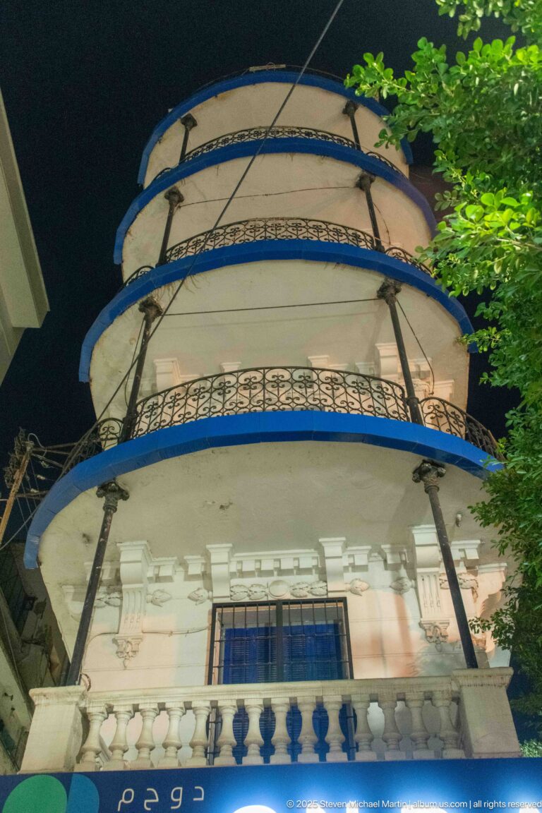 Rounded white apartment façade at night with stacked wrought-iron balconies, blue trim, balustrades, and cornice detail.