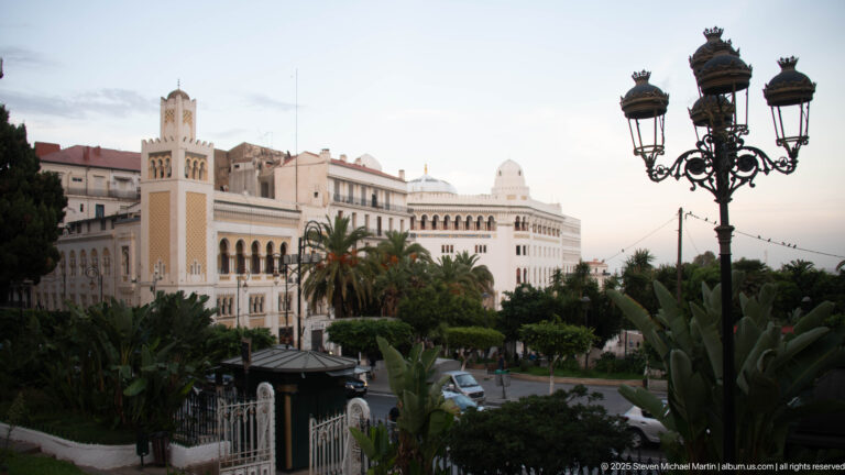 Palm-filled formal garden facing ornate white buildings with a square tower, domes, and decorative streetlamps.