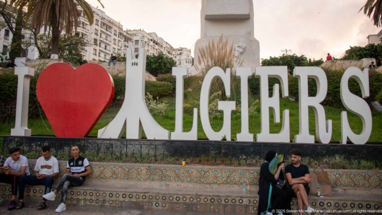 People seated beneath the large I heart ALGIERS sign in a landscaped public plaza.