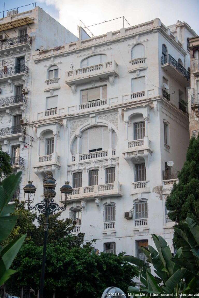 Large white apartment building with projecting bays, balconies, arched openings, and decorative moldings behind greenery.