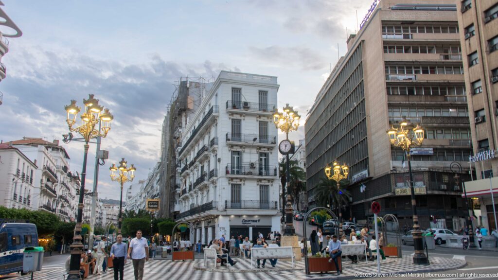 Busy Algiers boulevard with pedestrians, café seating, ornate streetlights, historic façades, and a modern office block.