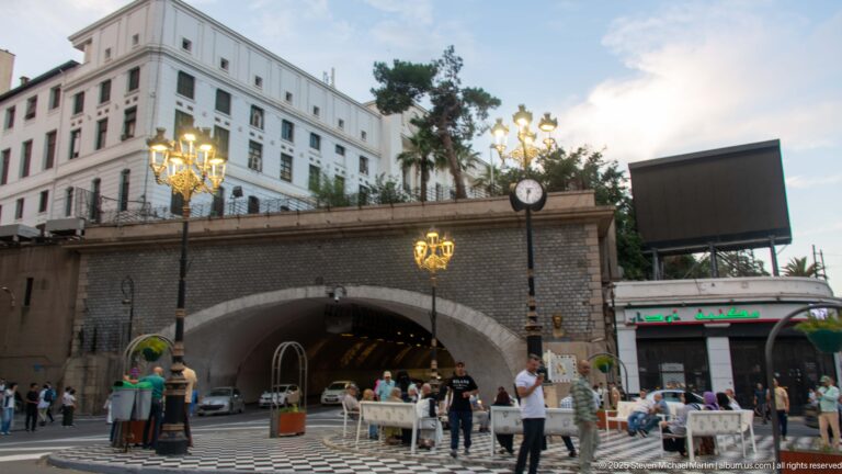 Stone-lined urban arch carrying a landscaped terrace above pedestrians and vehicles, with ornate lamps and a street clock.