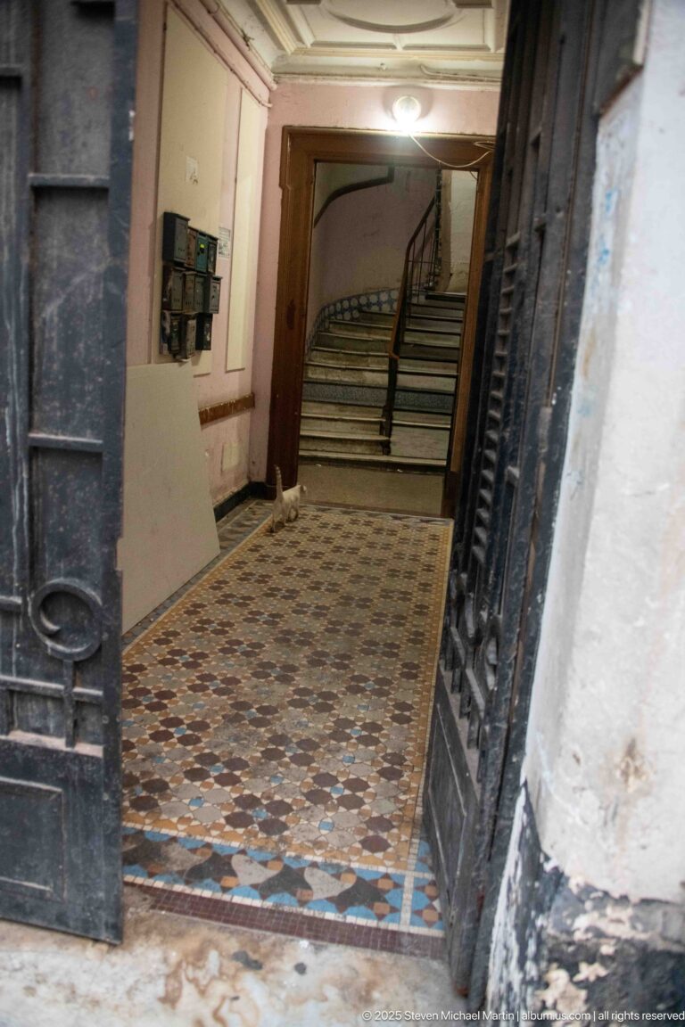 Open metal doorway leading into a tiled residential vestibule with electrical boxes and a staircase at the rear.