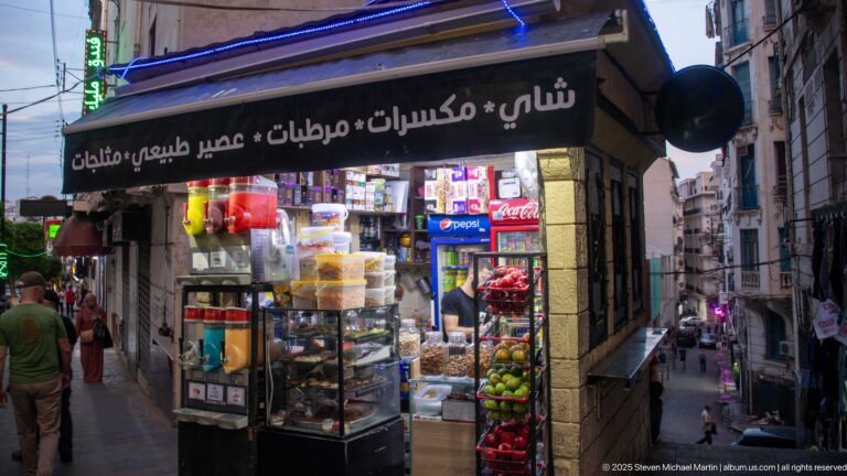 Small brightly lit shop with Arabic signage, drinks, packaged foods, fruit, and customers on a sloping Algiers street.