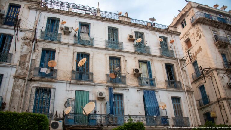 Weathered white apartment building with blue shutters, balconies, air conditioners, and many satellite dishes.