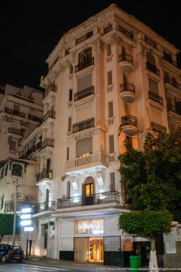 White multi-story building at night with balconies, dark railings, and a brightly illuminated street-level entrance.