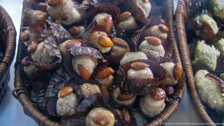 Basket of glossy bite-size sweets in paper cups, many topped with whole almonds and dark reddish-brown coatings.