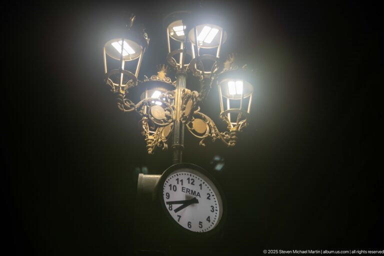 Ornate streetlight with multiple glowing lanterns, decorative metal scrollwork, and a round analog clock hanging below.