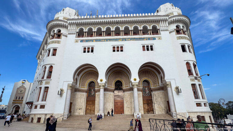 Large white building with three arched portals, upper arcade, corner towers, and broad entrance steps in Algiers.
