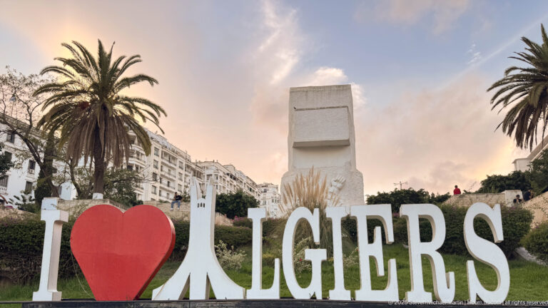 Large I heart ALGIERS sign in a landscaped plaza with palms and a tall pale monument behind it.
