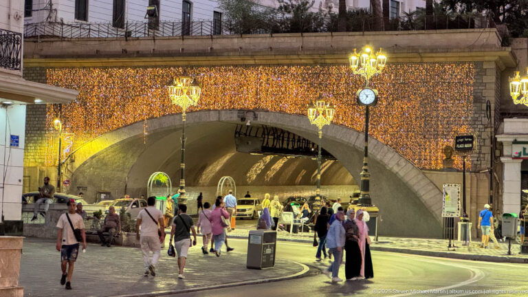 Pedestrians entering a brightly illuminated arched underpass at night beneath decorative streetlights and a clock.