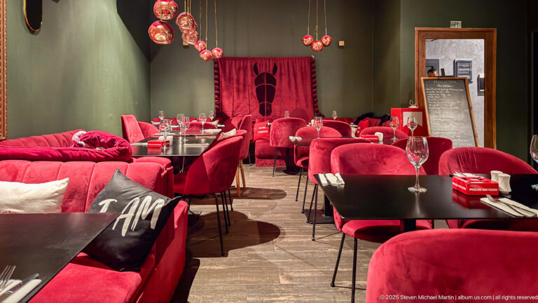 Restaurant interior with red velvet chairs and sofas, black tables, glassware, and hanging red decorative lights.