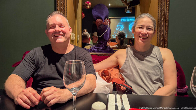 Two smiling diners seated at a restaurant table with a wine glass and colorful decorative interior behind them.