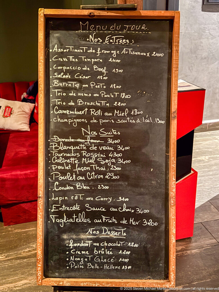 Black chalkboard with a handwritten French restaurant menu beside red seating in Algiers.