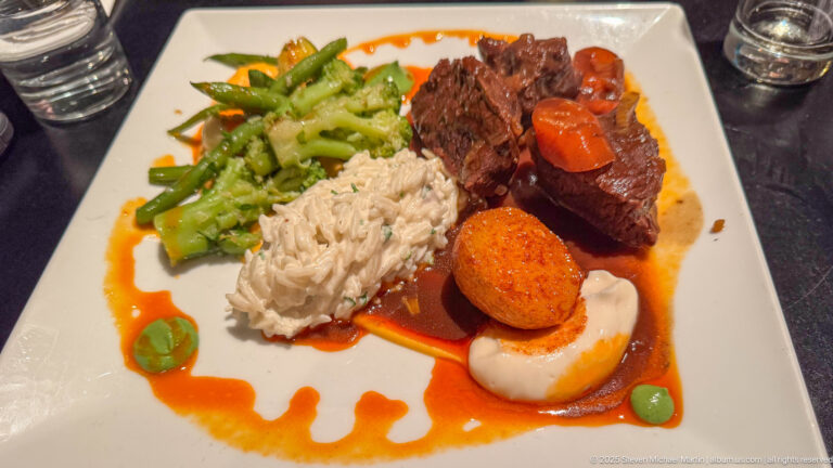 Plated entrée with braised meat, rice, green vegetables, carrots, a potato croquette, and red-orange sauce.