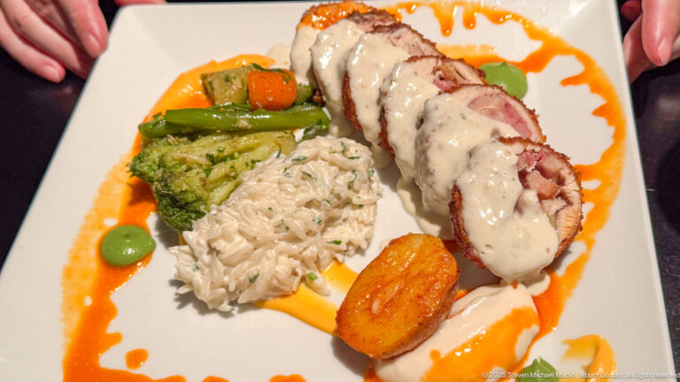 Sliced rolled meat with cream sauce, rice, broccoli, vegetables, and a browned potato portion on a decorated plate.