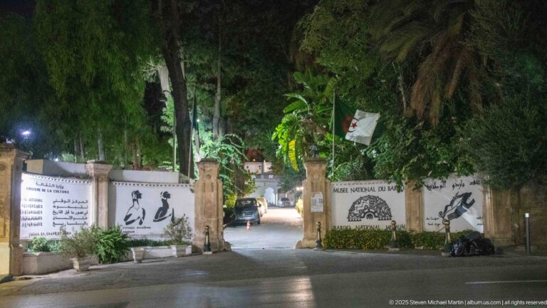 Night entrance to the Musée National du Bardo with wall signage, palm trees, an Algerian flag, and a gated drive.
