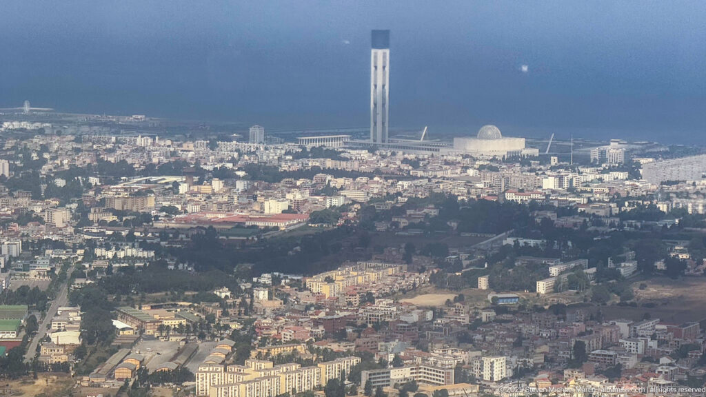 Elevated Algiers cityscape with dense neighborhoods and the tall minaret and dome of the Great Mosque near the coast.