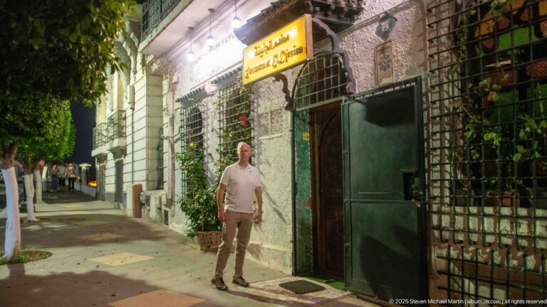 Man standing outside an Algiers restaurant at night beside a green door, metal grilles, plants, and illuminated signage.