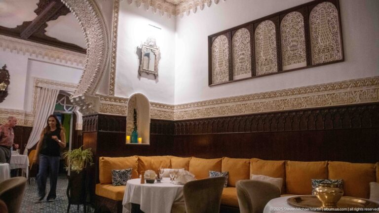Ornate dining room with carved dark wood, white patterned plasterwork, pierced panels, mustard banquette, tables, and standing guests.