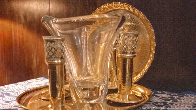 Clear glass pitcher and drinking glass with two metal vessels arranged on a round brass-toned tray against dark wood.