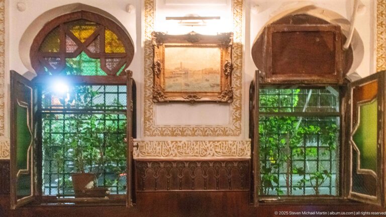 Two decorated windows with dark carved wood, patterned wall ornament, framed artwork, colored glass, and green foliage outside.