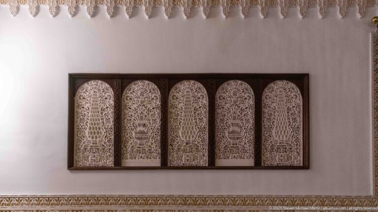 Dark framed panel with five narrow arched sections of intricate pierced ornament mounted on a white wall.
