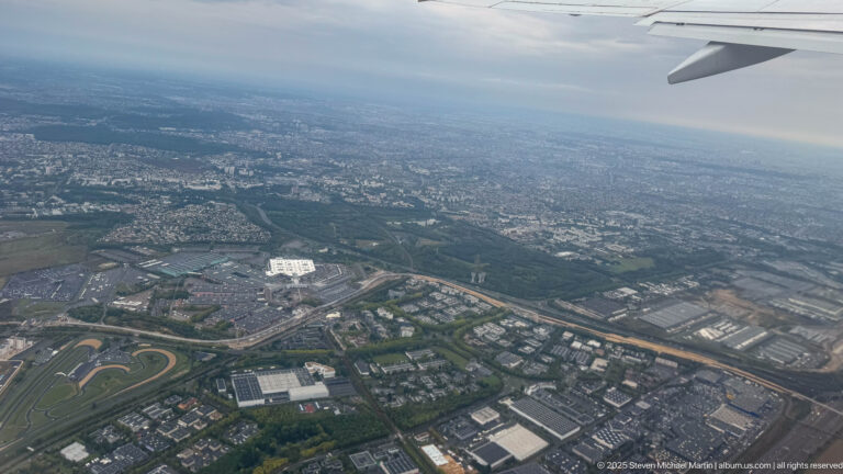 Oblique aerial view from an airplane showing dense urban development, roads, large buildings, and an oval racetrack below.