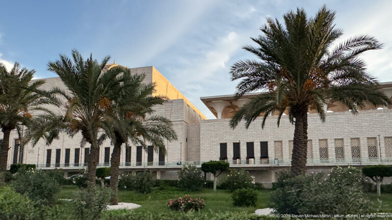 White modern institutional building with vertical window bays, angular roofline, palm trees, shrubs, and landscaped gardens.