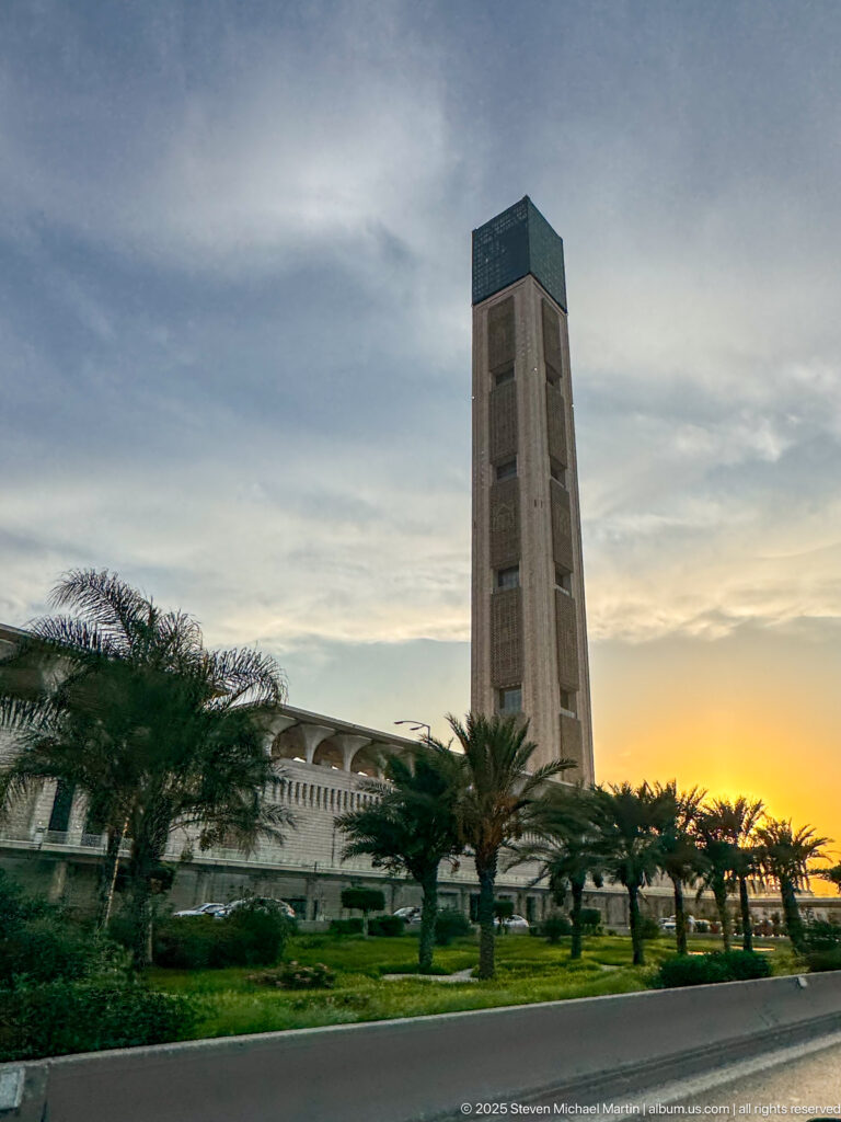 Tall rectangular tower above a low palm-lined complex and landscaped grounds at sunset in Algiers.